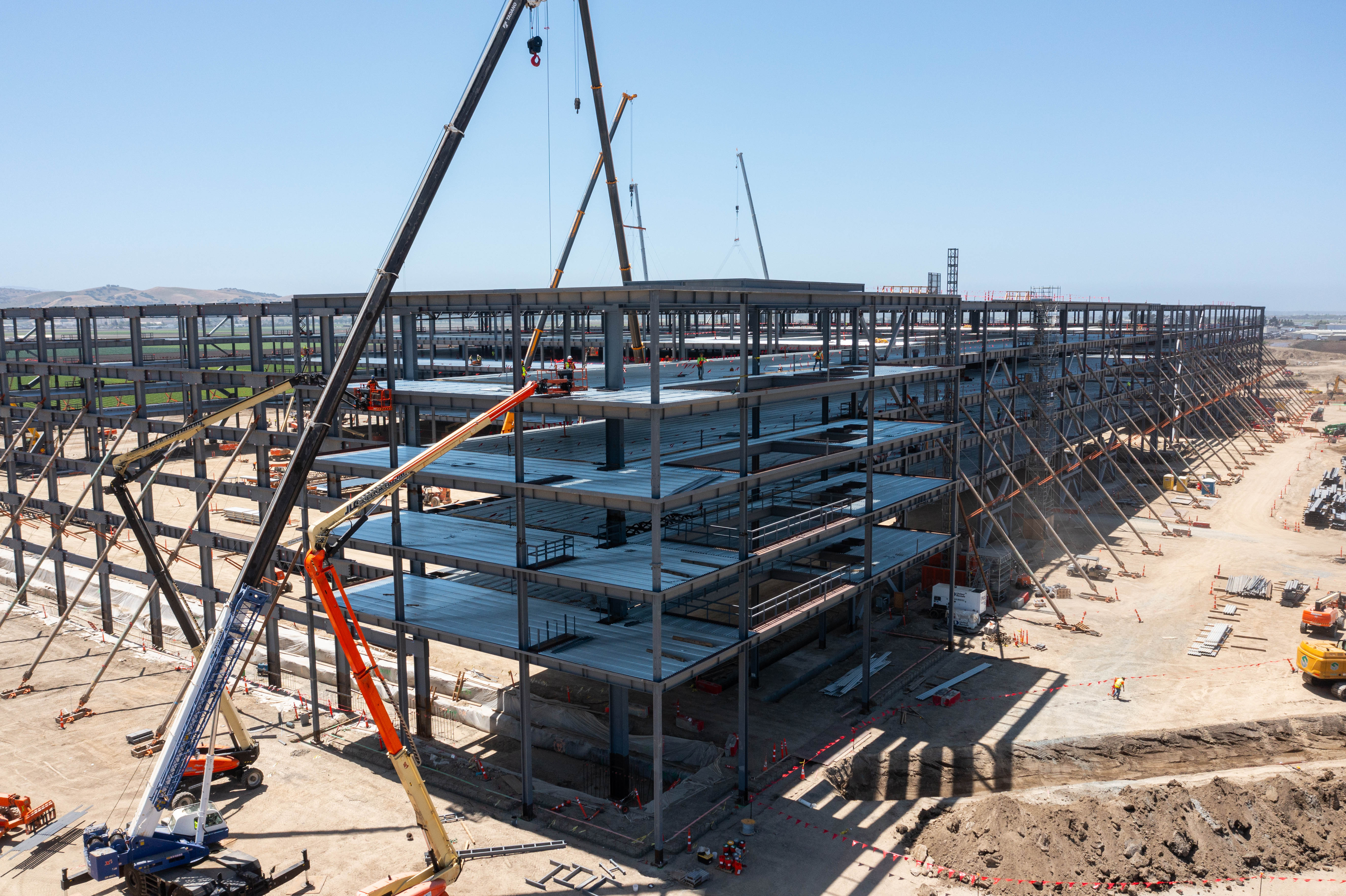 Multi-story steel frame building under construction with cranes and construction equipment on site in a large development area.