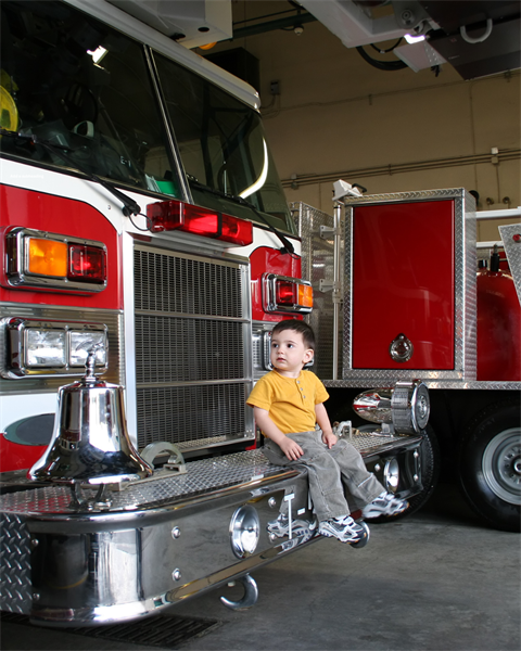 A red fire truck with chrome front and grille inside a garage, with a small child in a yellow shirt standing near the front bumper and looking toward the camera.
