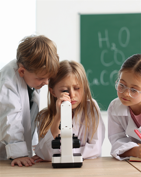 stock image of Three children in lab coats gathered around a microscope on a table