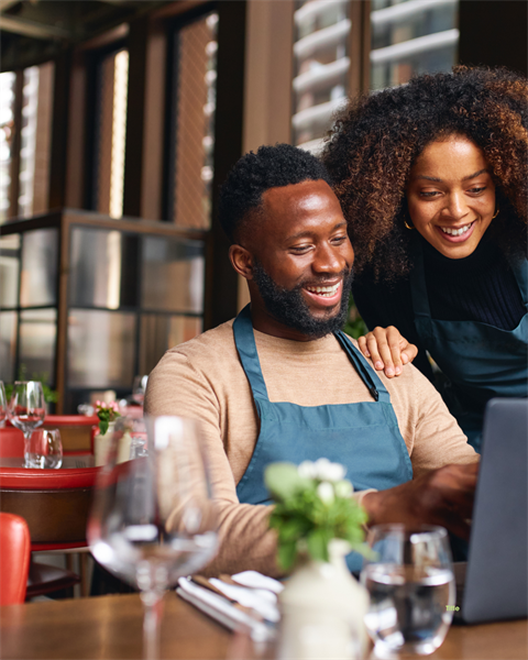Stock Image of two restaurant workers, one man and one woman, smile while looking at a laptop in a restaurant setting.