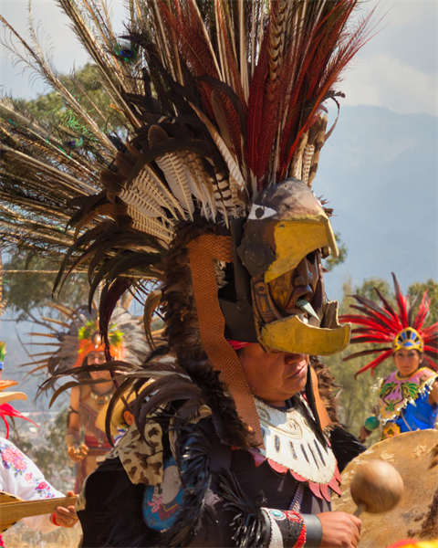 An Aztec dancer in an eagle warrior helmet and a massive feathered headdress plays a traditional drum at an outdoor cultural gathering.