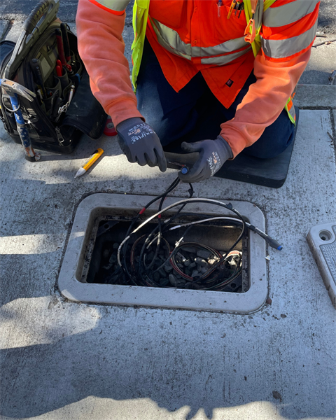 PA City of Salinas worker wearing an orange shirt, safety vest, and gloves kneels on a sidewalk to repair electrical wiring inside an open ground utility box.