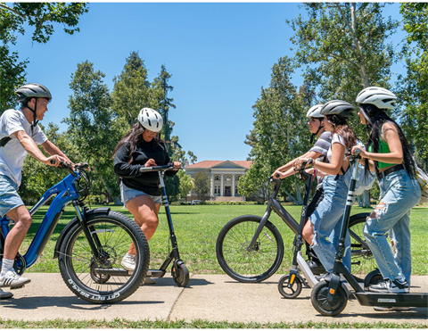 Five young adults with helmets pose on electric bikes and scooters on a park path, with a large lawn and classical building in the background.
