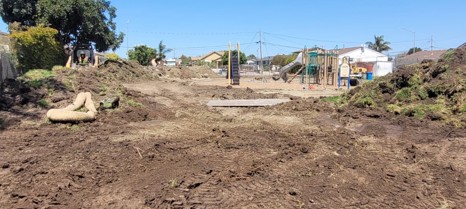 A dirt construction site with a cleared roadbed, scattered rocks and soil, and distant playground equipment and buildings along the street under a blue sky.