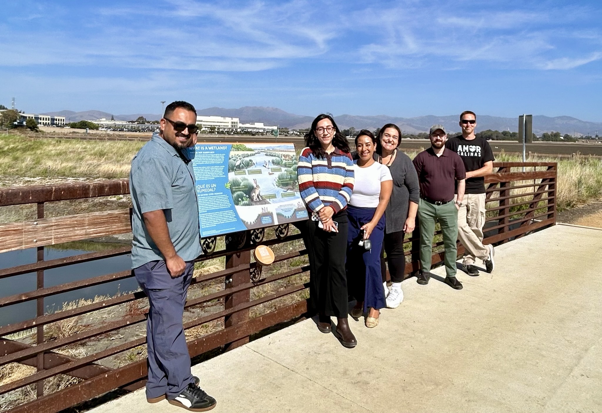 A group of six people stand outdoors on a sunny path beside a wooden fence, posing with a large blue informational sign and a view of a landscape in the background.