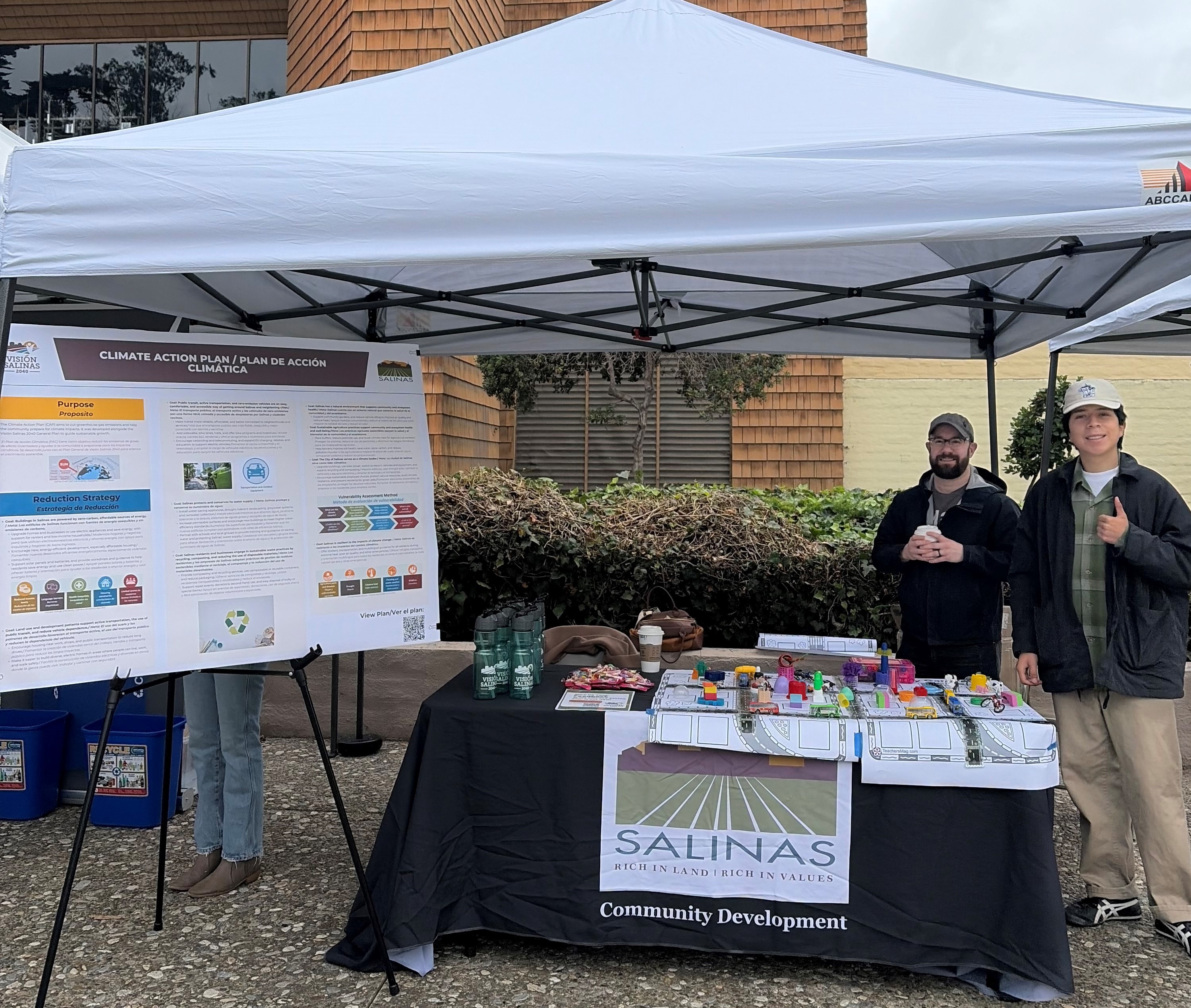 Outdoor community event with four black-and-white canopy tents; a table with brochures and a sign reading “SALINAS” in front, and two people standing near a display board in a courtyard between brick buildings.