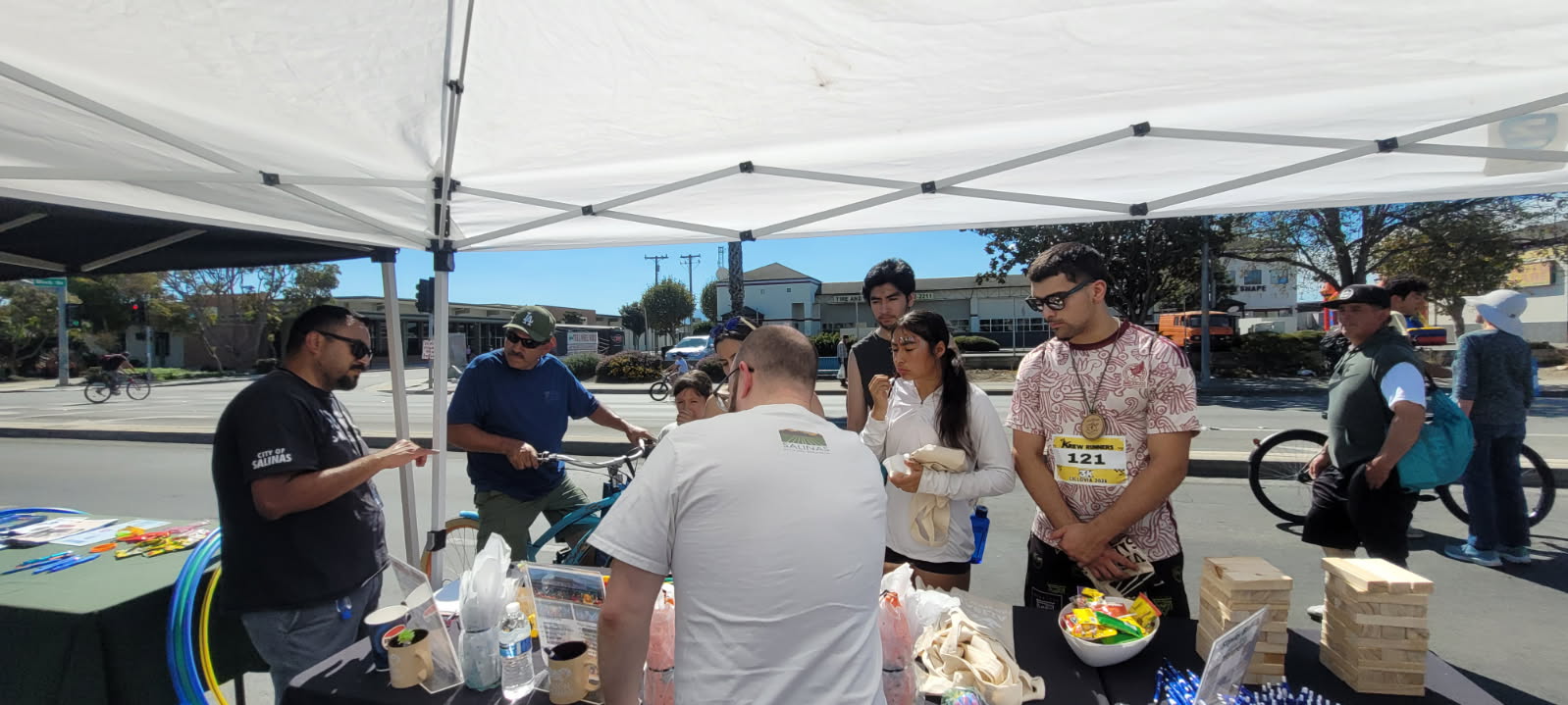 People gather under an outreach tent at an outdoor local street event. People on bikes, standing and chatting with items on a table for giveaways. 