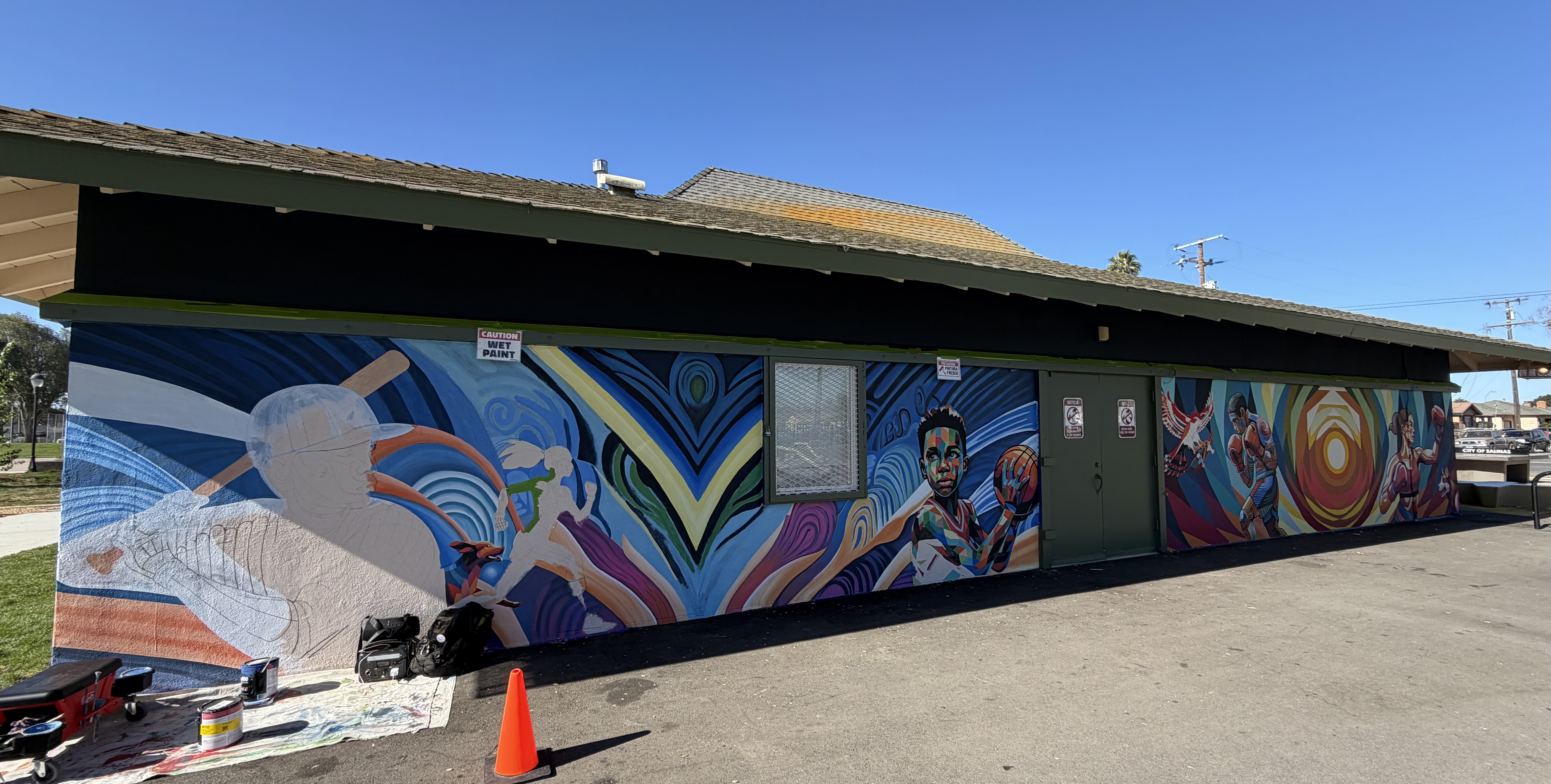 Wide view of a long building wall covered with colorful mural panels of people and figures, with a doorway, a dark roof, and a street in the foreground with an orange cone.