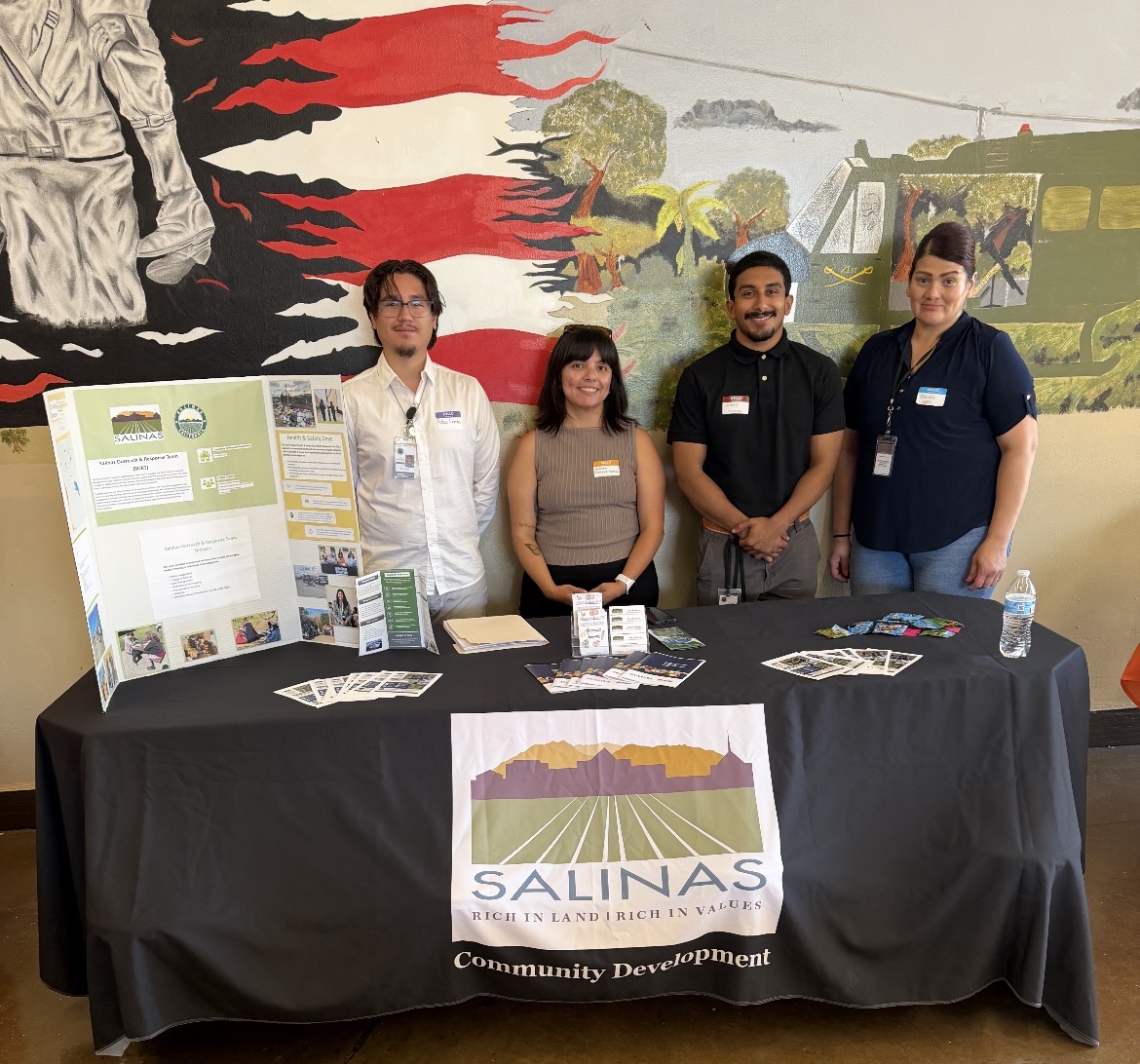 Four people standing behind an information table covered with a black tablecloth featuring the City of Salinas logo, which reads 