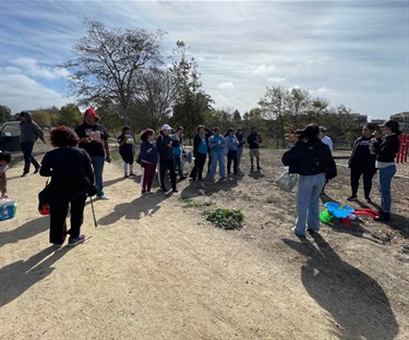  Gemini said A large group of people gathers on a dirt path and field near a playground under a bright, overcast sky. Many individuals are dressed in casual athletic wear or matching blue t-shirts, while a colorful toy wheelbarrow sits on the ground in the foreground.